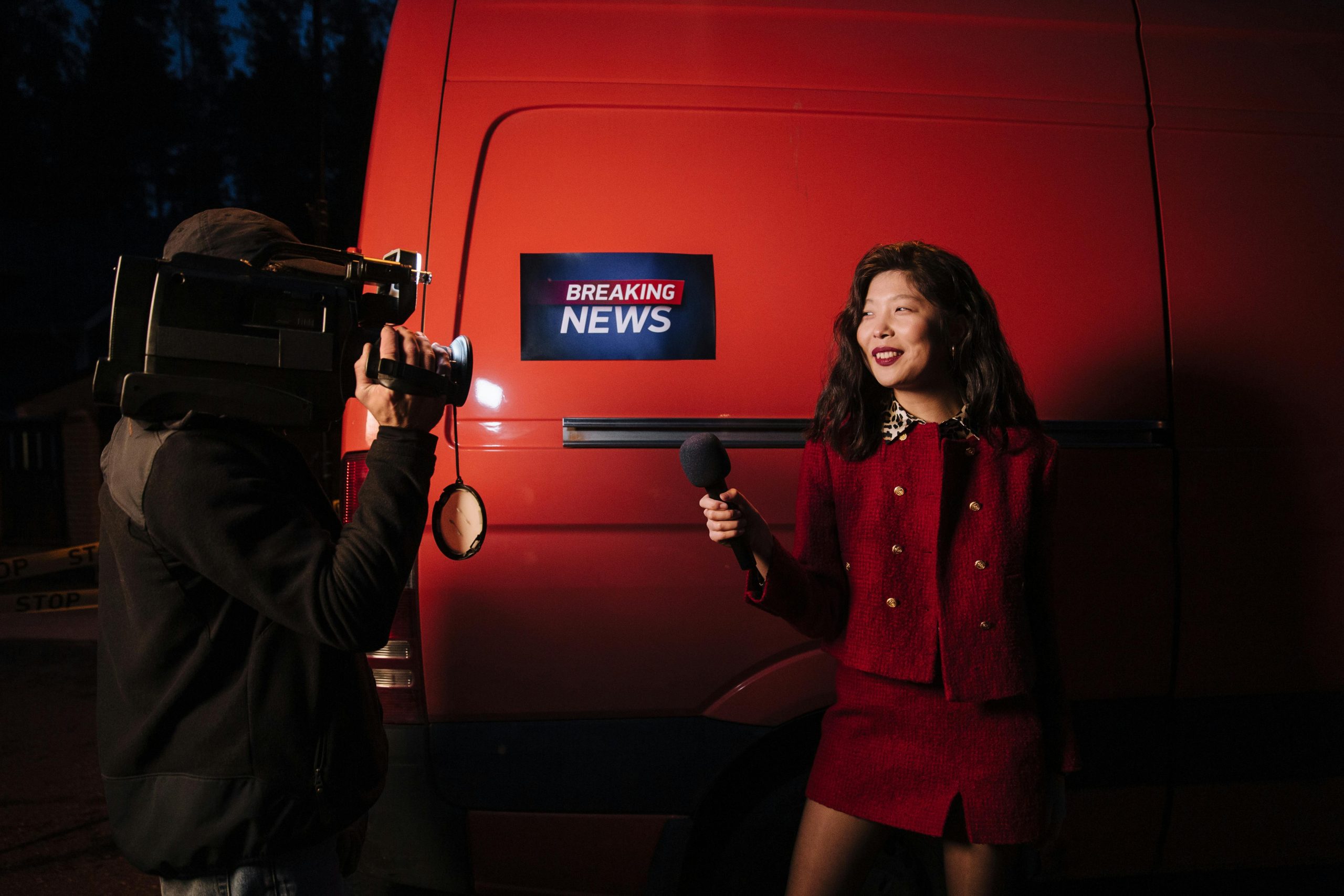 Dynamic news reporter holding microphone near red van during nighttime outdoor broadcast.